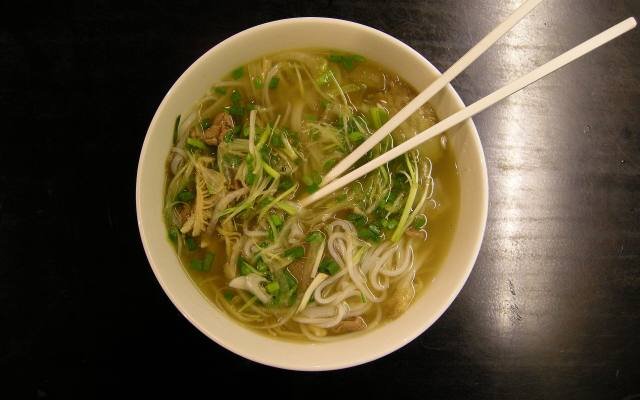 A bowl of pho with rice noodles, herbs and chopsticks on a dark table